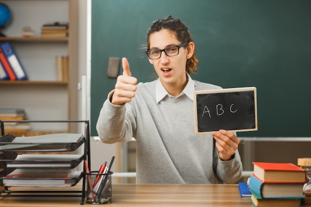 写真 教室で学校の道具をつけて机に座っているミニ黒板を持って眼鏡をかけている若い男性教師に親指を立てて笑っている