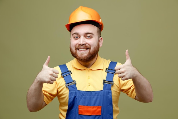 Фото Улыбающийся молодой строитель в униформе, показывающий большие пальцы на зеленом фоне