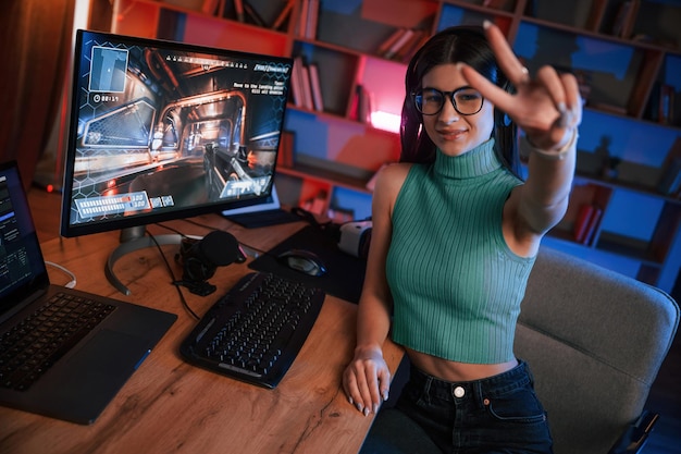 Photo smiling showing victory gesture young pretty female gamer is playing shooter neon lighting