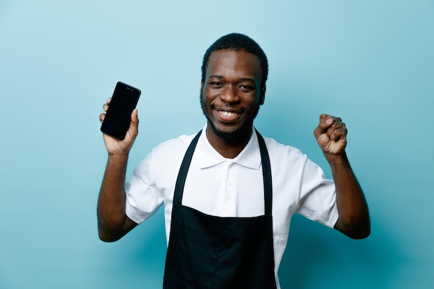 Smiling showing yes gesture young african american barber in uniform holding phone isolated on blue background