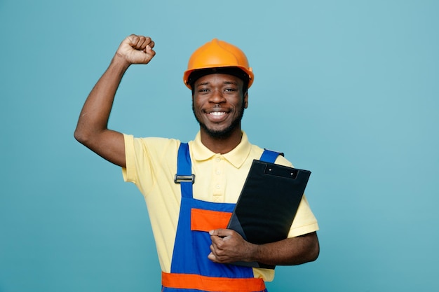 Sorridente mostrando sì gesto giovane costruttore afroamericano in uniforme che tiene appunti isolato su sfondo blu