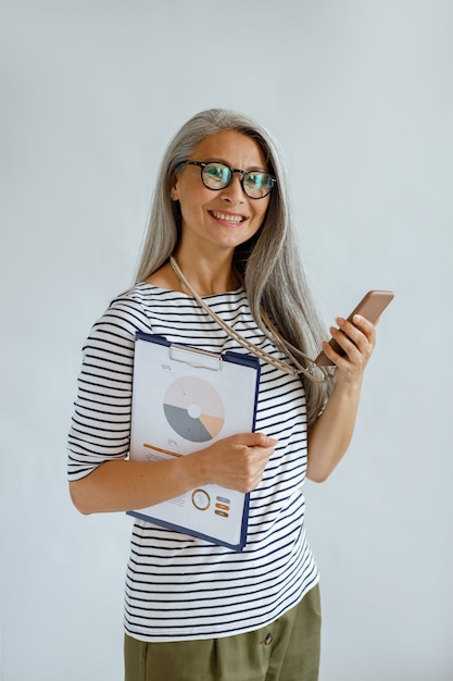 Foto sorridente signora asiatica dai capelli d'argento con gli occhiali tiene diagrammi colorati e smartphone in piedi su sfondo grigio chiaro in studio