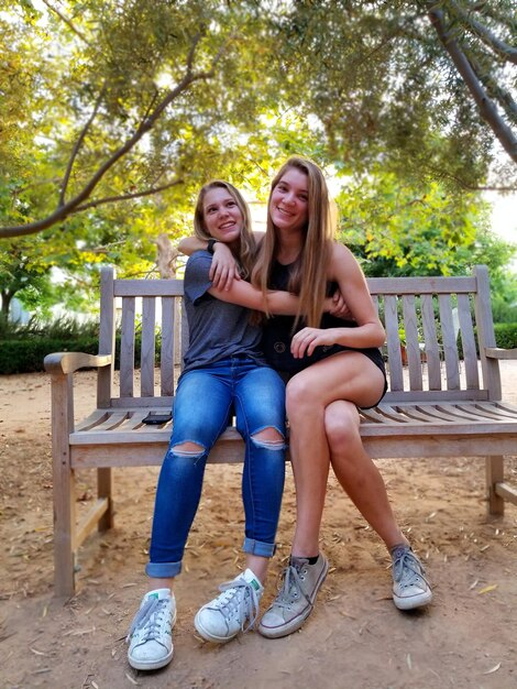 Foto sorelle sorridenti che si abbracciano mentre sono sedute su una panchina nel parco