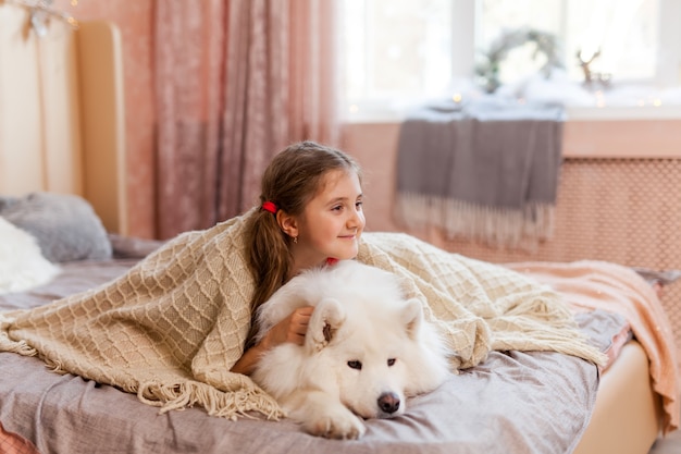 Smiling sleepy cute little girl hugging big Samoyed dog at home