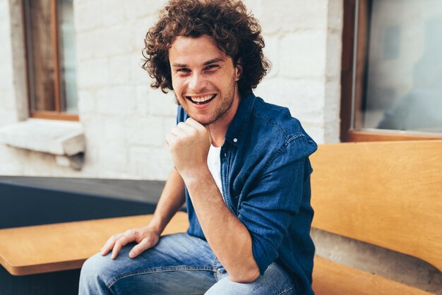 Smiling smart handsome young man with curly hair looking to the camera and sitting on the bench in the city street Happy male student relaxing outdoors People and lifestyle concept