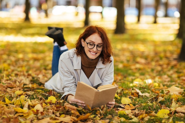 Foto sorridente intelligente piuttosto giovane donna europea in occhiali e impermeabile legge il libro si trova su foglie gialle nel parco