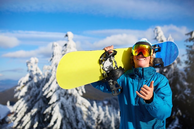 Smiling snowboarder posing carrying snowboard on shoulders at ski resort near forest before freeride