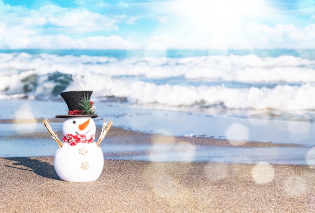 太陽が降り注ぐビーチで笑顔の雪だるま。メリークリスマスと幸せな新年のカードの休日の概念