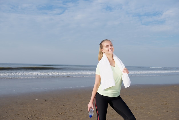 Smiling sports woman standing with water bottle at the beach