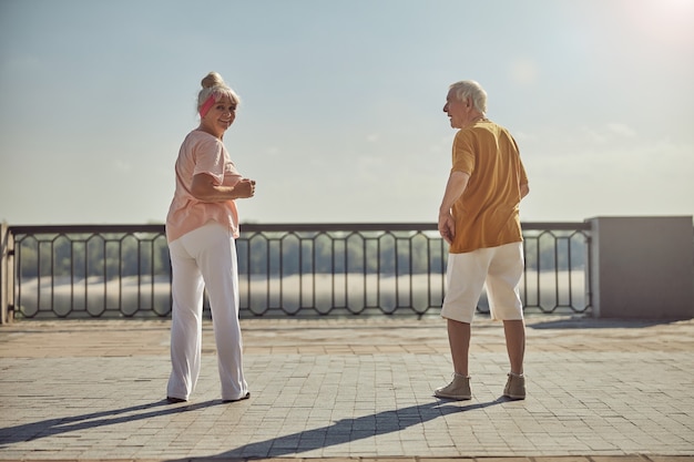 Smiling sporty aged female dressed in casual clothes working out with a gray-headed man outside