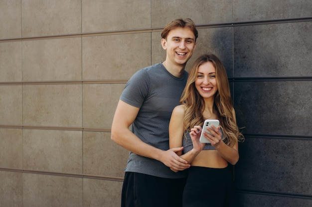 Smiling sporty couple using mobile phone on gray wall