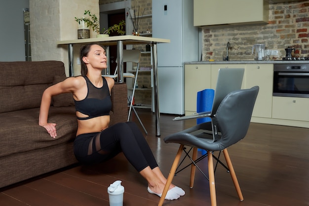 A smiling sporty girl in a black tight suit is doing triceps and chest workouts and watching an online training video on a laptop. A female coach conducting a remote fitness class at home.