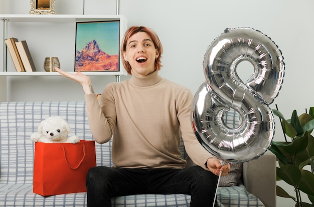 Smiling spreading hand handsome guy on happy women day holding number eight balloon sitting on sofa in living room