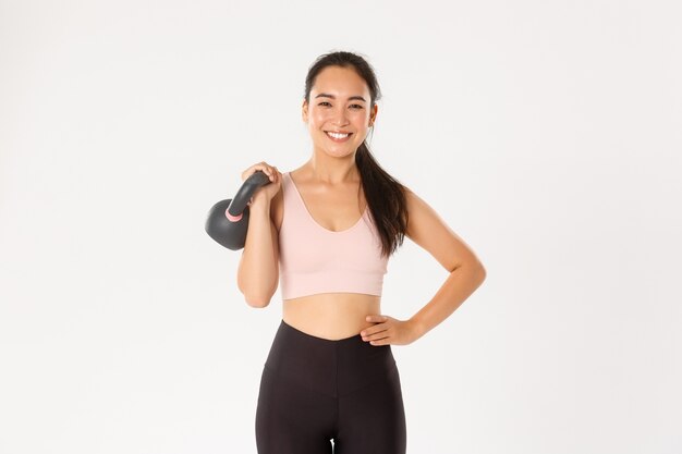   Smiling strong and slim asian fitness girl, female athlete holding kettlebell and looking carefree, gaining muscles at gym, standing  