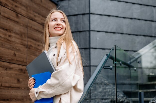 Фото Улыбающаяся девушка студент держит папки, тетради, книги в руках улыбается, смотрит в сторону на фоне современного здания университета. копировать пространство