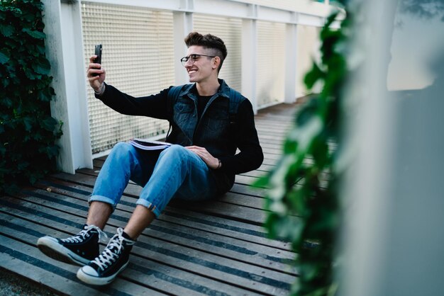 Smiling student in glasses taking selfie while sitting on floor