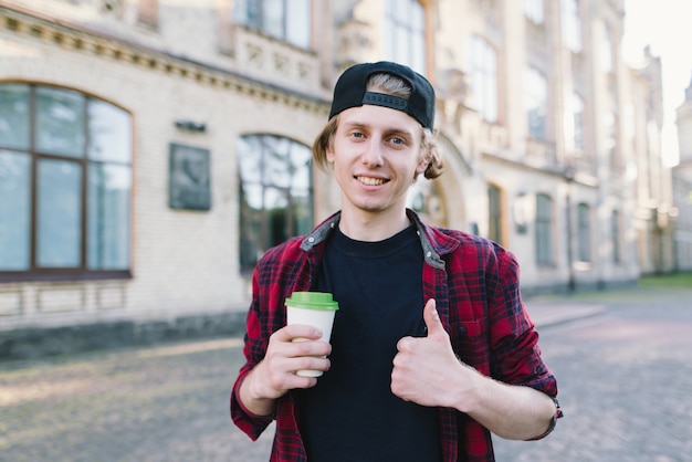 Улыбающийся студент держит кофе и показывает палец вверх против университета.