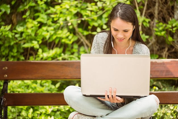 Smiling student sitting on bench listening music and using laptop