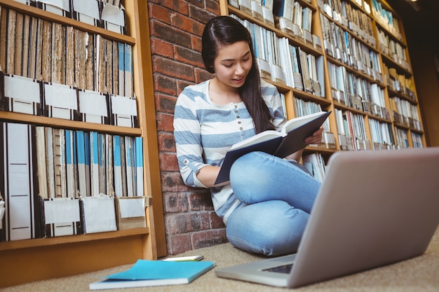 Smiling student sitting on the floor against wall in library studying with laptop and books