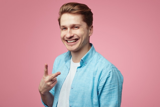 Smiling student wearing blue shirt and is gesturing vsign over pink wall