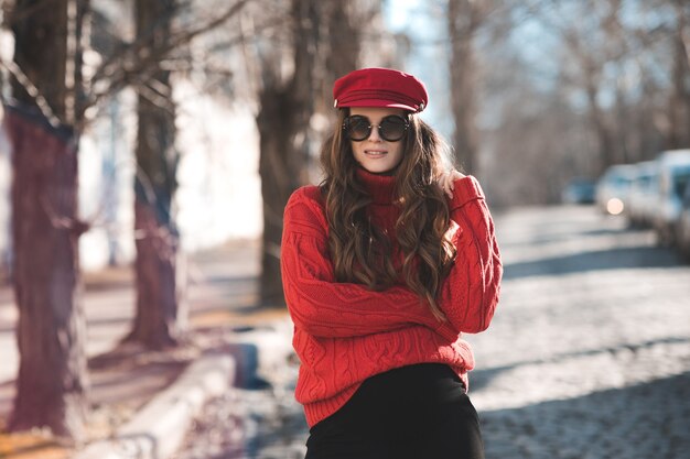 Sorridente donna elegante che indossa un maglione lavorato a maglia rosso e un cappello con occhiali da sole