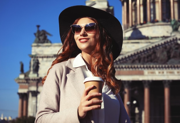 Smiling stylish young woman drinking coffee while walking on a city street