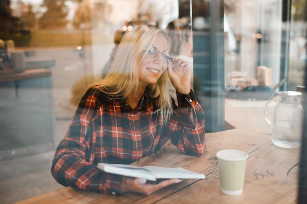 Smiling stylish young woman wear glasses and reading paper book in cafe through window close up