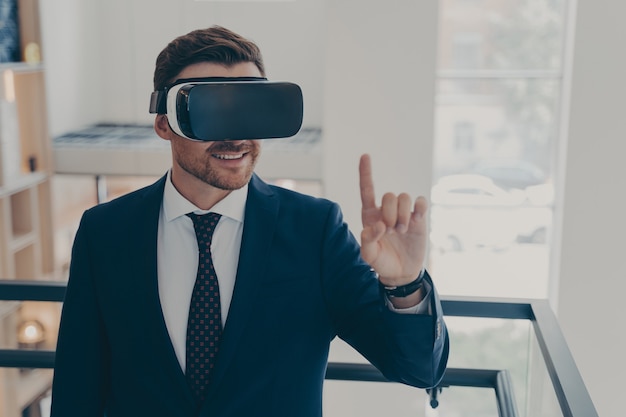 Smiling successful businessman in suit standing in office interior and using VR glasses for work