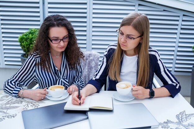 Smiling successful female workers are working together, working on a laptop on the terrace, female colleagues are busy discussing ideas, girls in a cafe at a business meeting. Business concept