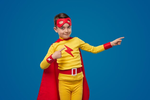 Smiling superhero boy pointing away in studio