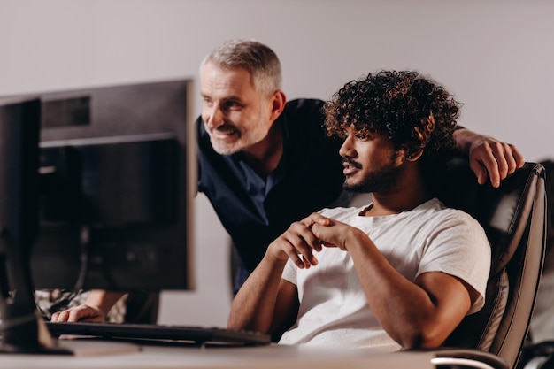 Photo smiling supervisor looking at his employee's computer older colleague helping young office worker