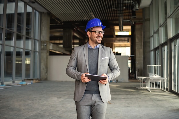 Photo smiling supervisor walking in building in construction process with tablet in hands and checking on works