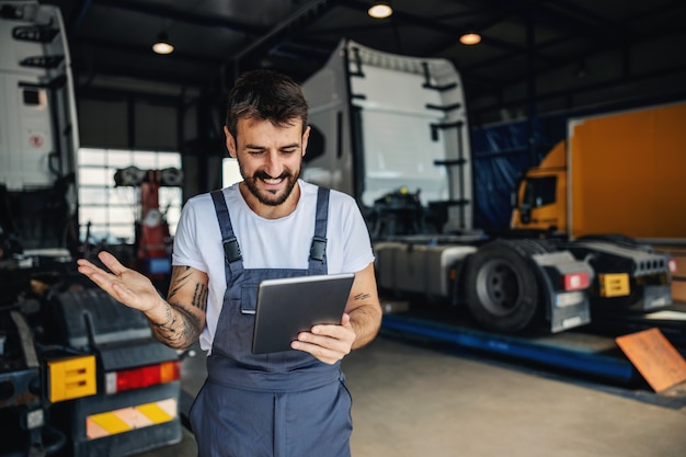 Smiling tattooed bearded blue collar worker in overalls using tablet to check on delivery while standing in garage of import and export firm