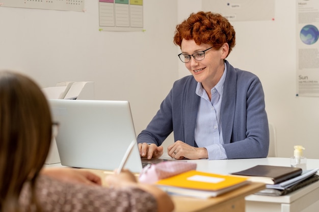 Smiling teacher in eyeglasses using laptop by workplace