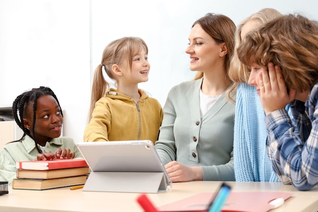 Smiling teacher using digital tablet while talking with pupils in classroom Education learning