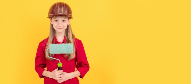 Smiling teen child in builder hard hat with paint roller on yellow wall Child builder in helmet horizontal poster design Banner header copy space