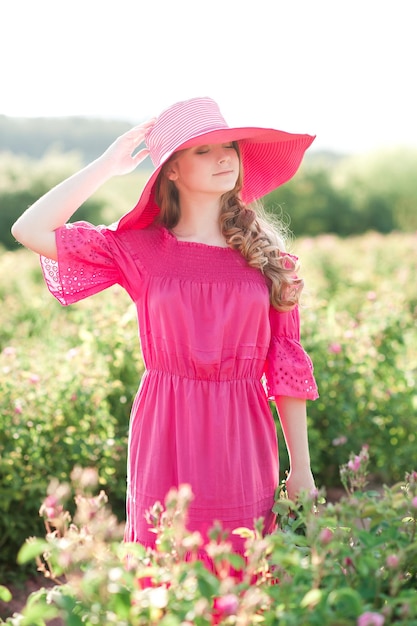 Foto ragazza adolescente sorridente di 14-16 anni che indossa un vestito rosa e un cappello in piedi in un campo di rose in estate