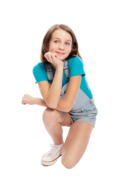 Smiling teen girl in denim overalls squats. Isolated on a white background