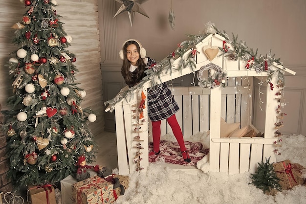 Улыбающаяся девочка-подросток в ушанках веселится в украшенном рождественском домике возле новогодней елки, готовится к рождественскому празднику, празднованию приветствия