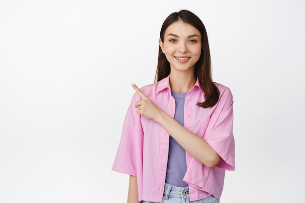 Smiling teen girl pointing finger at upper left corner showing sale banner place for advertisement standing in stylish outfit over white background