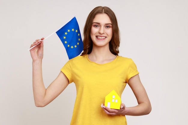 Улыбающаяся девушка-подросток, работница агентства недвижимости в желтой футболке, держащая бумажный игрушечный домик и флаг Европейского союза, смотрит в камеру с зубастой улыбкой. Внутренний студийный снимок изолирован на сером фоне