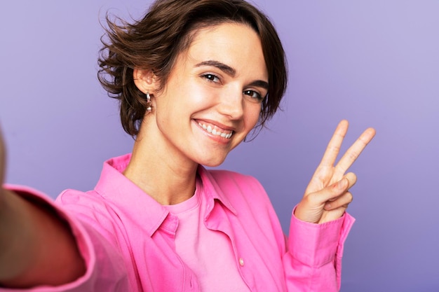 Foto ragazza adolescente sorridente che mostra un gesto di pace e si fa un selfie guardando la telecamera del telefono cellulare