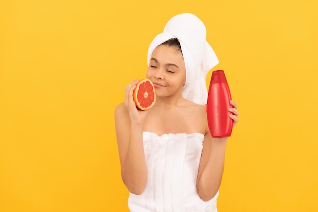 Smiling teen girl in towel with grapefruit shampoo bottle on yellow background