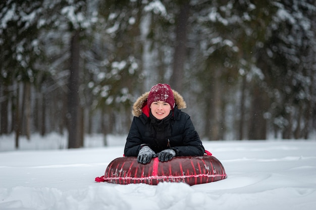 A smiling teenage boy in a black jacket with a fur and red hat lies on a tubing in a snowcovered for...