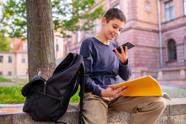 Smiling teenage boy seated under a deciduous tree with an open book and the cellphone