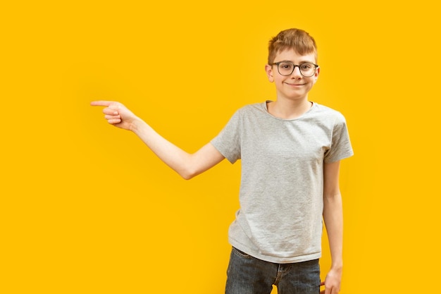 Smiling teenage boy wears glasses points with his index finger in the side Schoolboy on yellow background showing direction