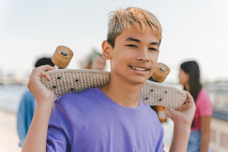 Photo スケートボードを握り,都市の通りでカメラを見つめている色の ⁇ の十代の少年が微笑んでいる