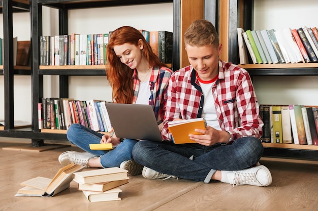 Sorridente coppia adolescente seduto su un pavimento presso lo scaffale per libri