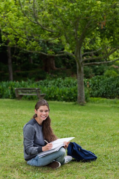 Улыбаясь девочка-подросток делает ее домашнее задание, сидя в парке