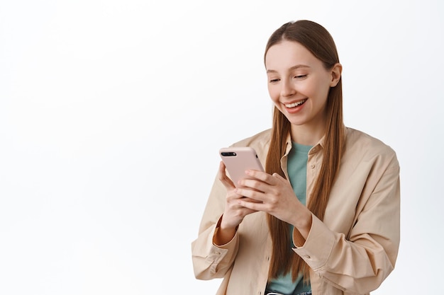 Smiling teenage girl, gen-z female student chatting on social media, reading phone screen, standing relaxed against white background